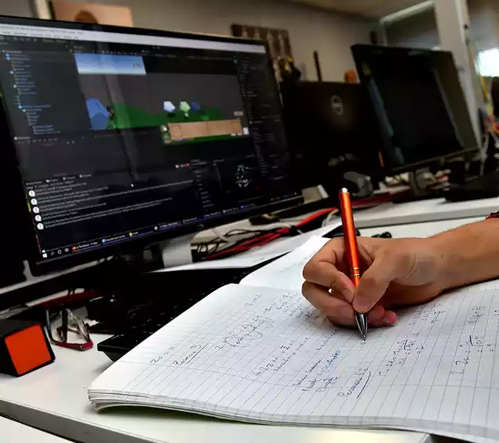 Cours de programmation Main d'un étudiant prenant des notes pendant un cours de programmation jeu vidéo à Creajeux, avec Unity affiché à l'écran.